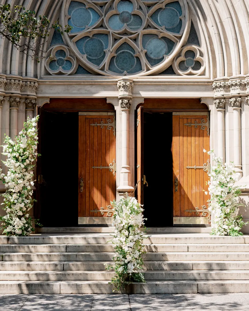 The carved wooden doors of Queen of All Saints church in Brooklyn, the stone steps and railings trailing white flowers.