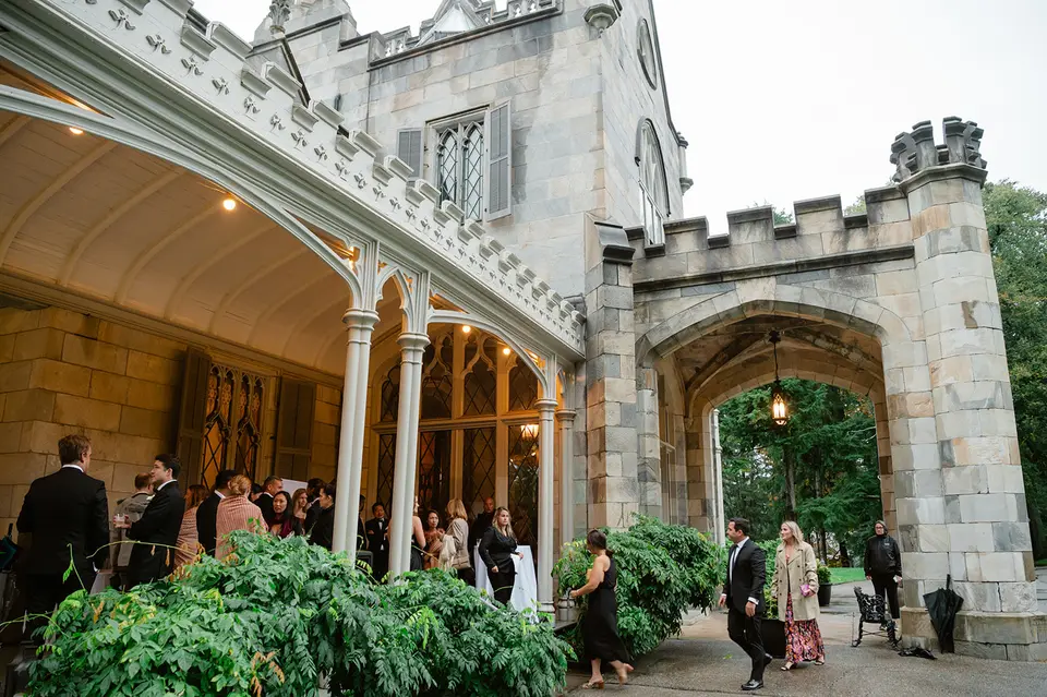 Guests gather on the mansion's lantern-lit porch beside a stone archway, the castle towers above them.