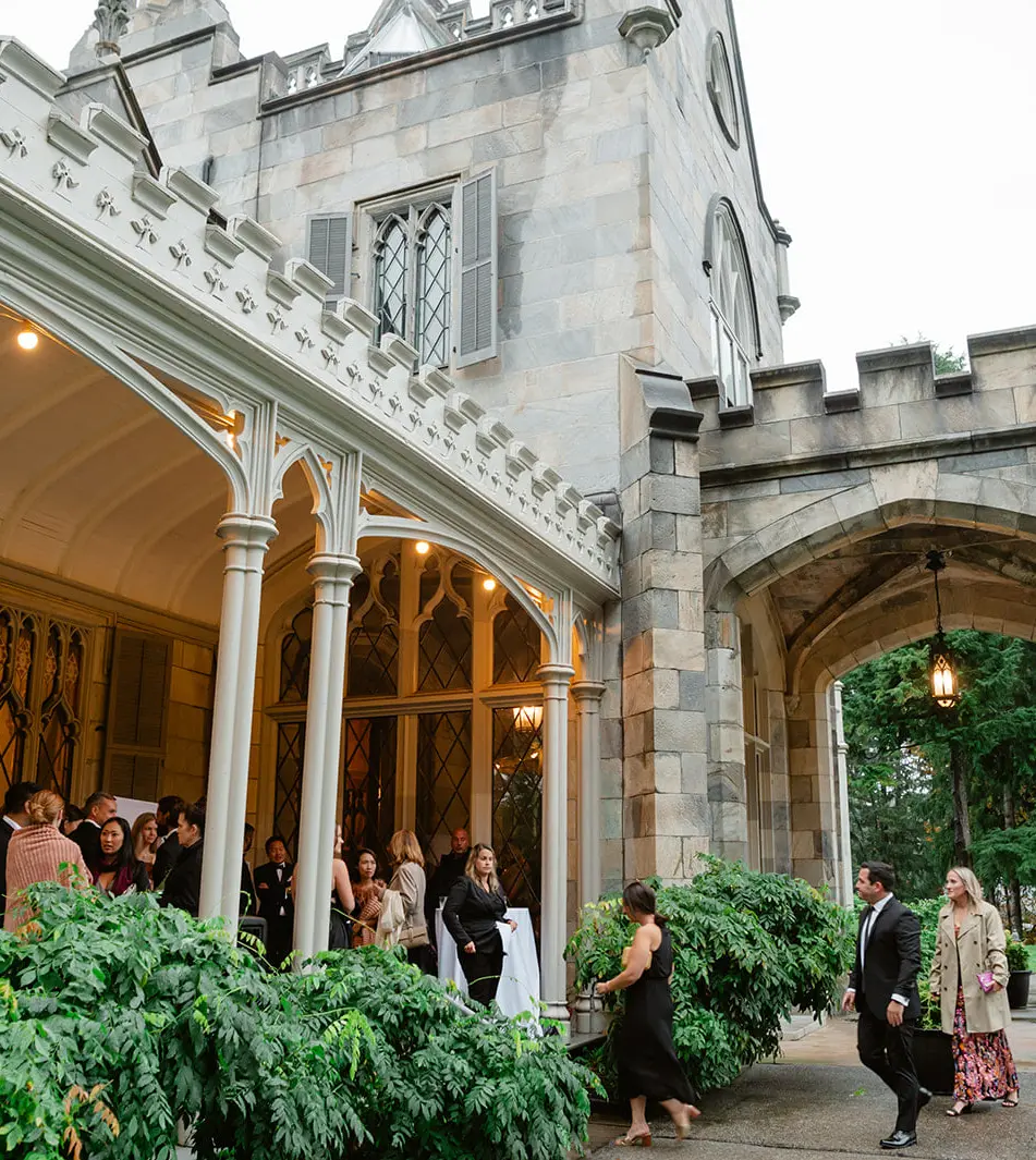 Guests gather on the mansion's lantern-lit porch beside a stone archway, the castle towers above them.