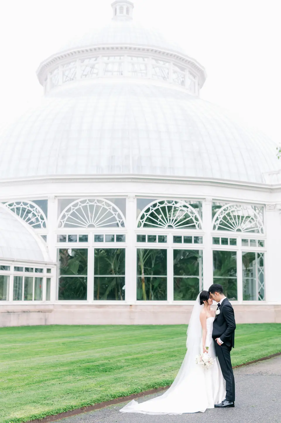 The couple kiss on the lawn before the white glass dome of the Enid A. Haupt Conservatory at The New York Botanical Garden.