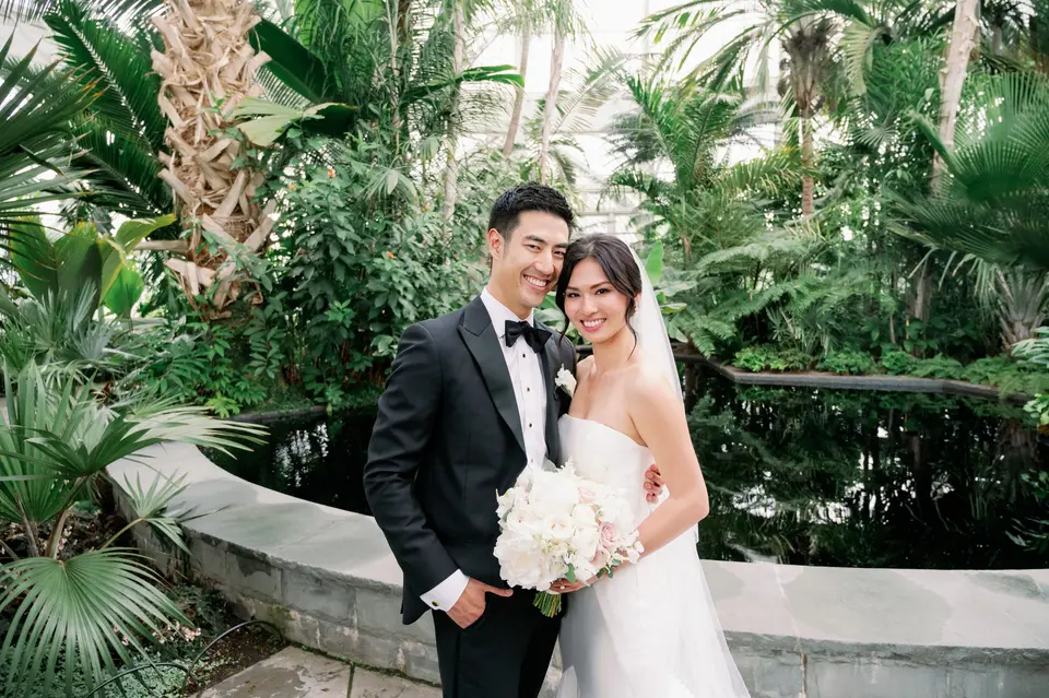 The couple stand smiling among tall palms and ferns inside the Victorian glasshouse.