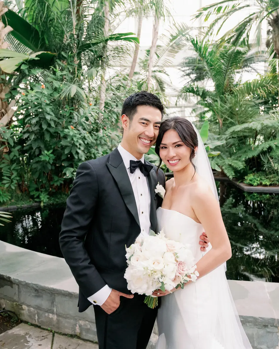 The couple stand smiling among tall palms and ferns inside the Victorian glasshouse.
