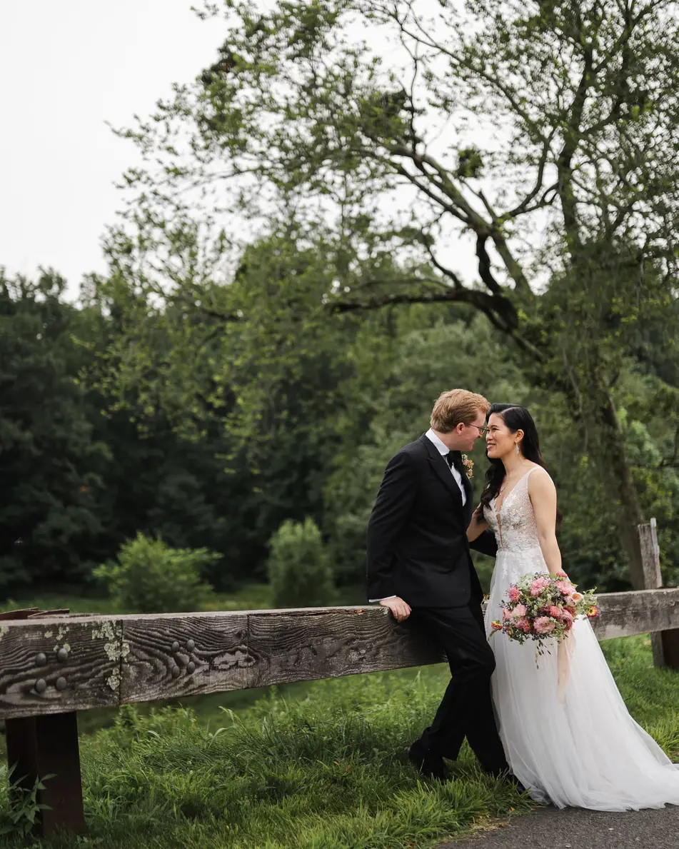The couple kiss by a wooden fence at the edge of green fields and trees on the farm.