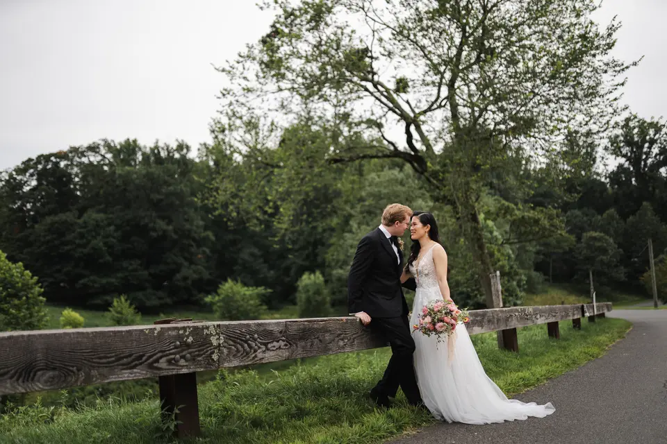 The couple kiss by a wooden fence at the edge of green fields and trees on the farm.