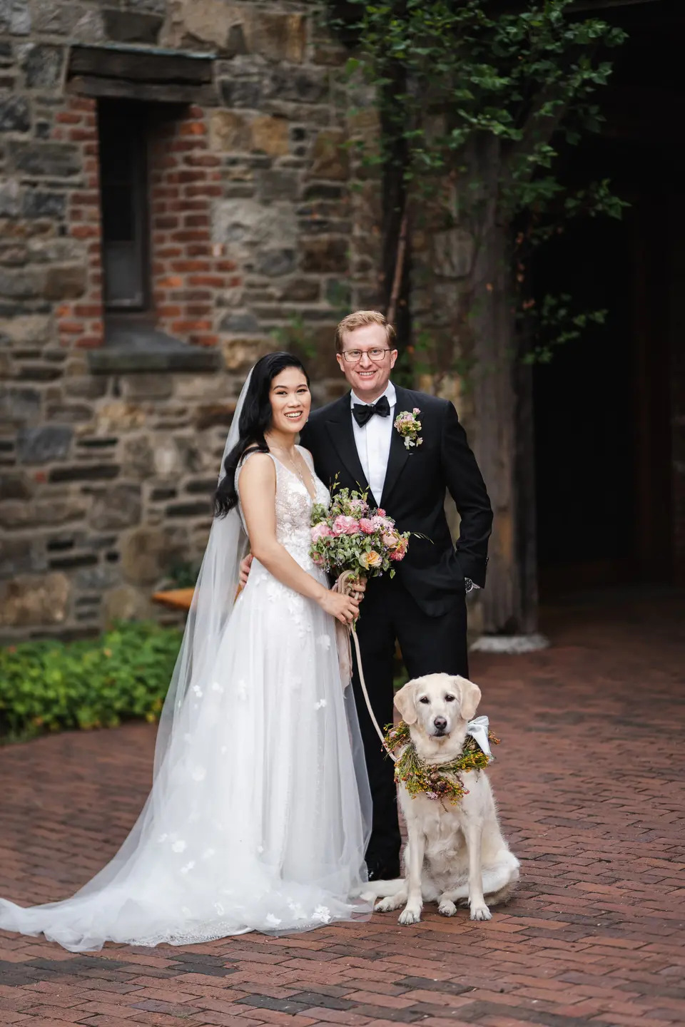 The couple stand by a stone wall with their golden retriever, who wears a wreath of flowers.