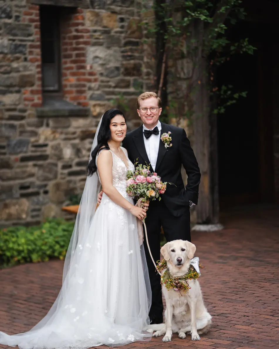 The couple stand by a stone wall with their golden retriever, who wears a wreath of flowers.