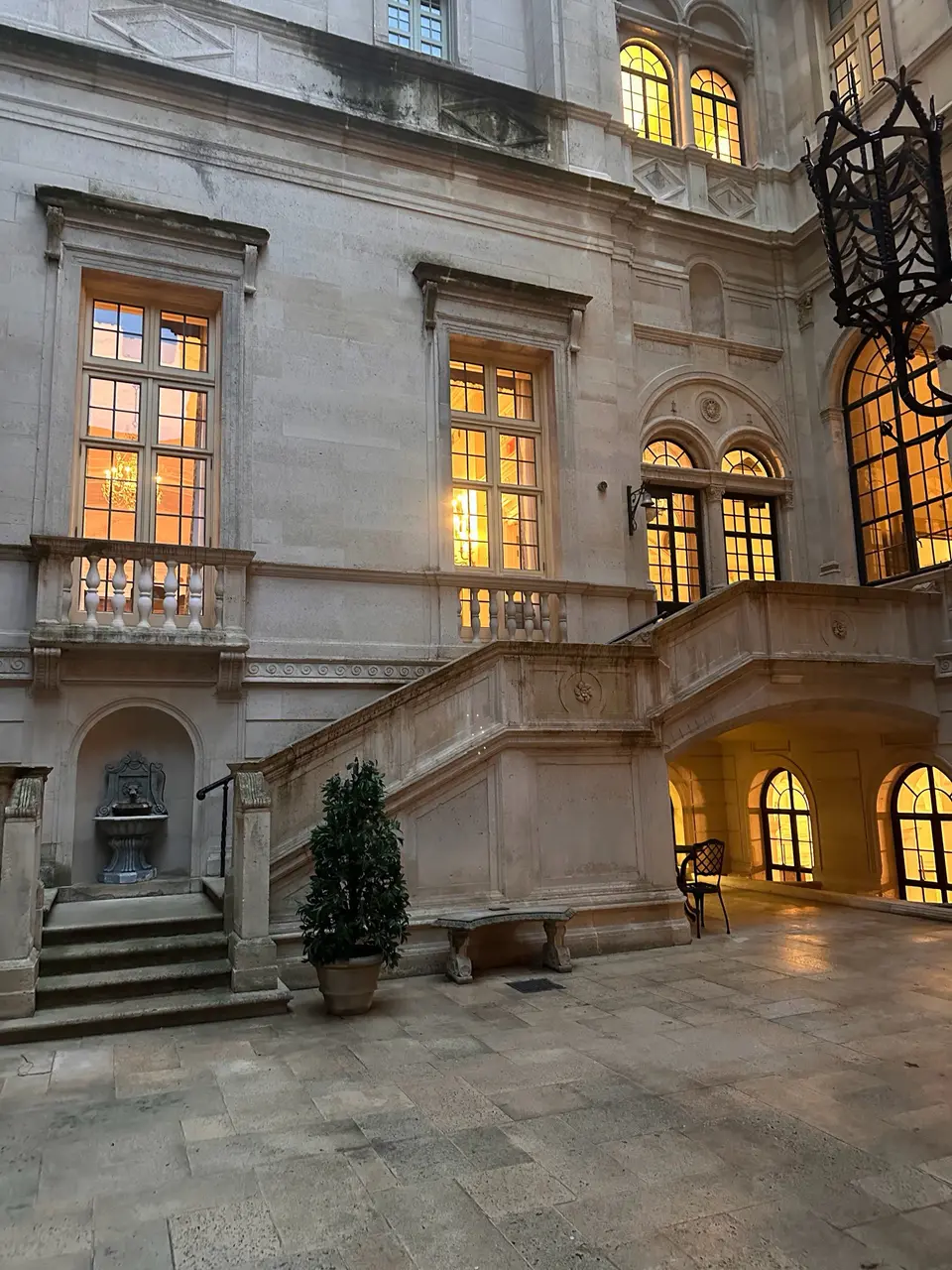 The stone courtyard of the Burden Kahn Mansions at dusk, its warmly lit windows, balustraded steps and carved facade rising around a potted tree.
