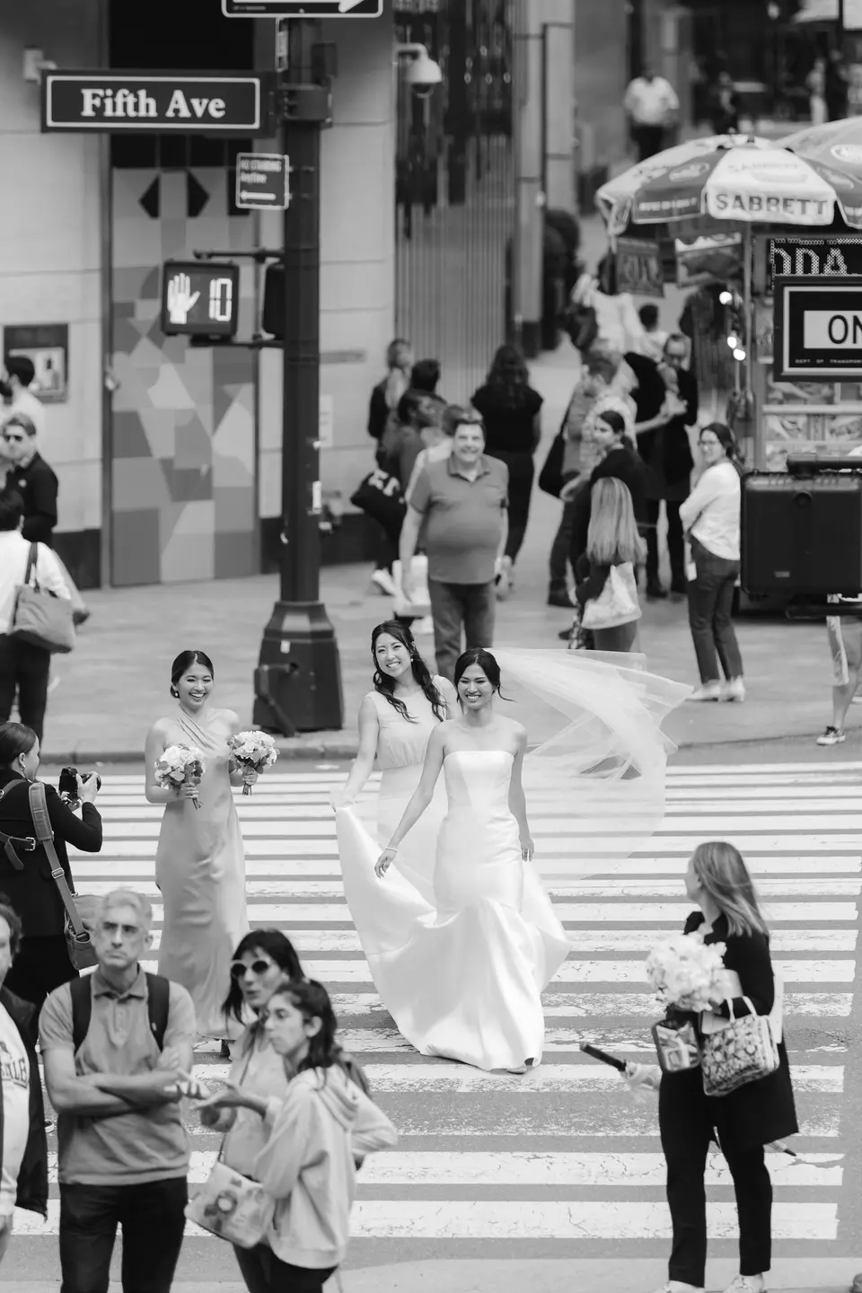 The bride crosses Fifth Avenue among New Yorkers, her veil lifting behind her. Black and white.