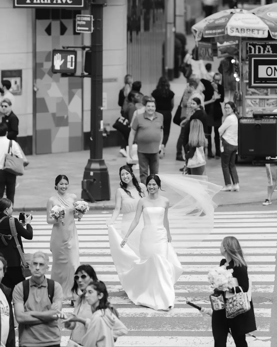 The bride crosses Fifth Avenue among New Yorkers, her veil lifting behind her. Black and white.