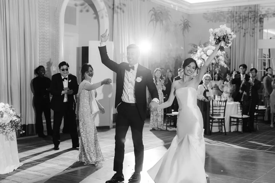 The couple dance into the room with their arms raised, guests clapping around them. Black and white.