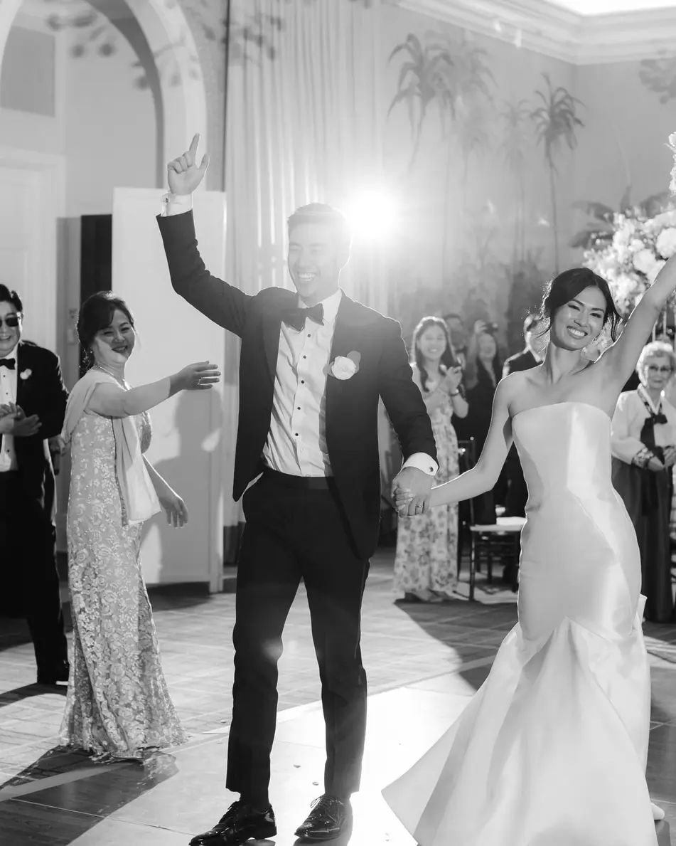 The couple dance into the room with their arms raised, guests clapping around them. Black and white.