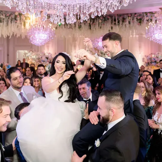 The bride is lifted high on a chair above the dance floor, guests crowding around her beneath the chandeliers.