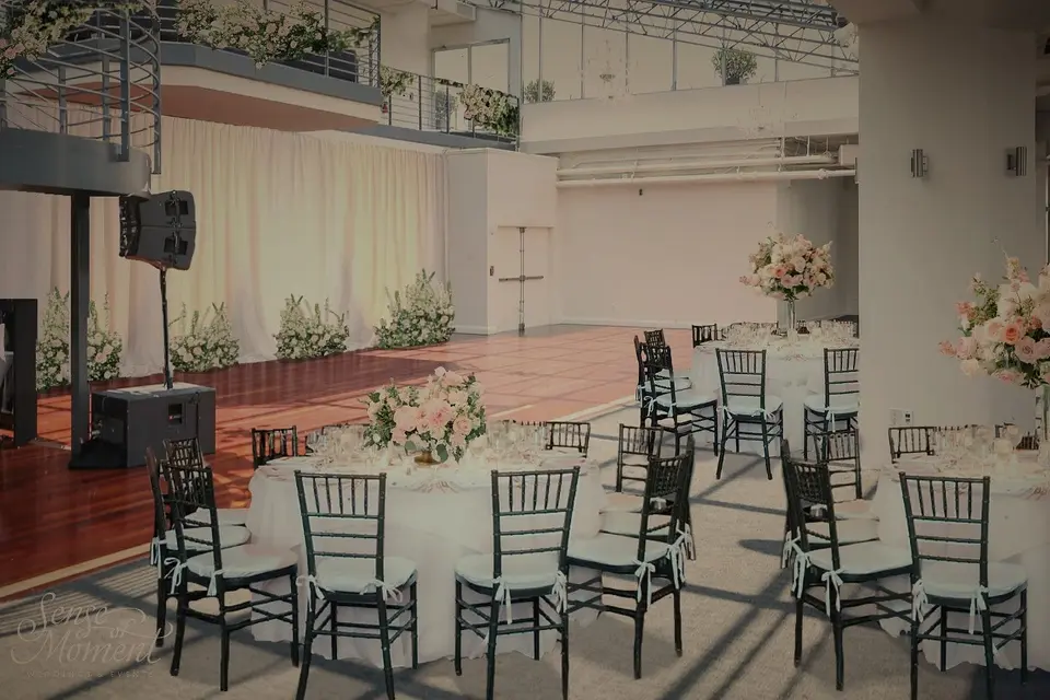A rendering of the reception room at Tribeca Rooftop, round tables set with low blush arrangements and black chairs, white drapes and low flowers behind a wood dance floor, under a glass roof.