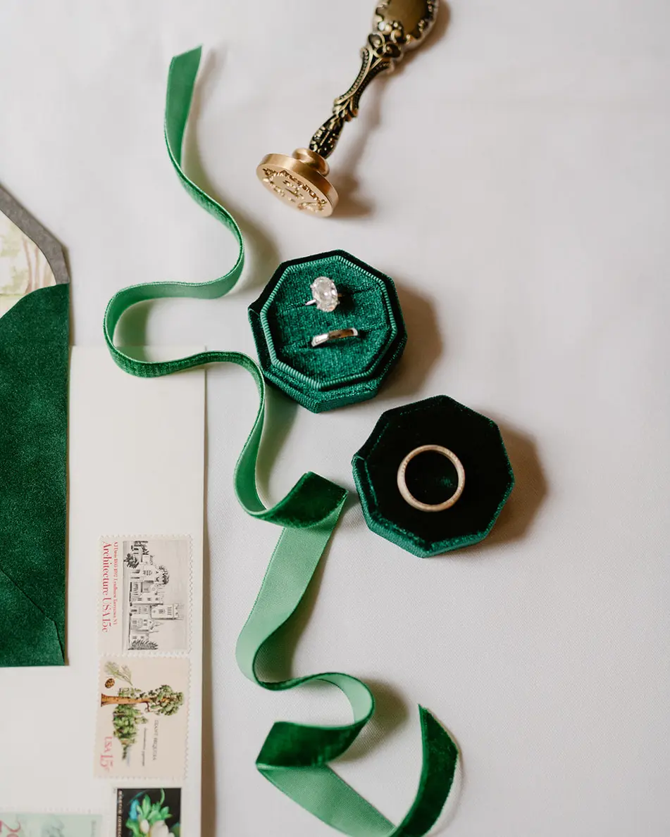 A flat lay of emerald velvet ribbon, two octagonal emerald ring boxes, a brass wax seal and the invitation suite on white.
