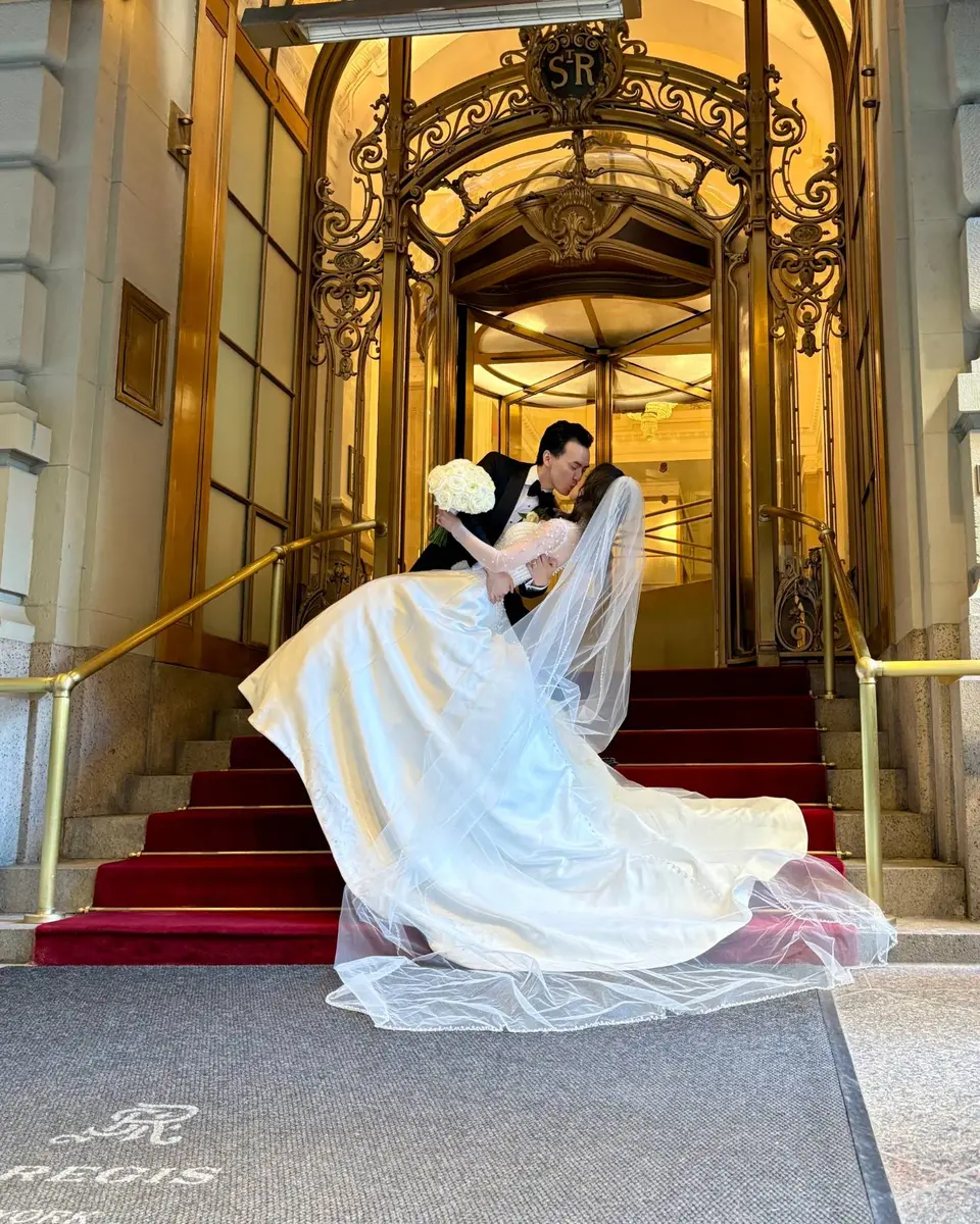 The groom dips the bride in a kiss on the red-carpeted steps of The St. Regis New York, her long veil and satin skirt sweeping across the stairs beneath the gilded revolving door.
