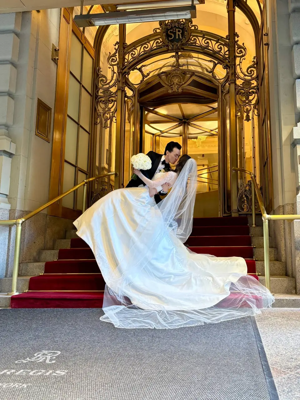 The groom dips the bride in a kiss on the red-carpeted steps of The St. Regis New York, her long veil and satin skirt sweeping across the stairs beneath the gilded revolving door.
