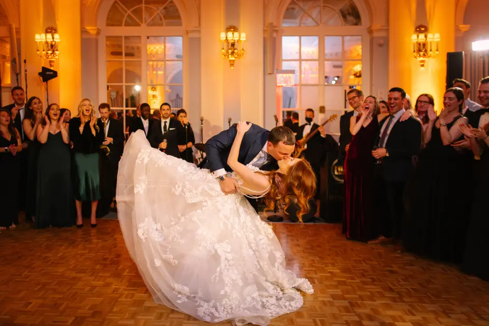 On the dance floor, the groom dips the bride low as guests cheer around them.
