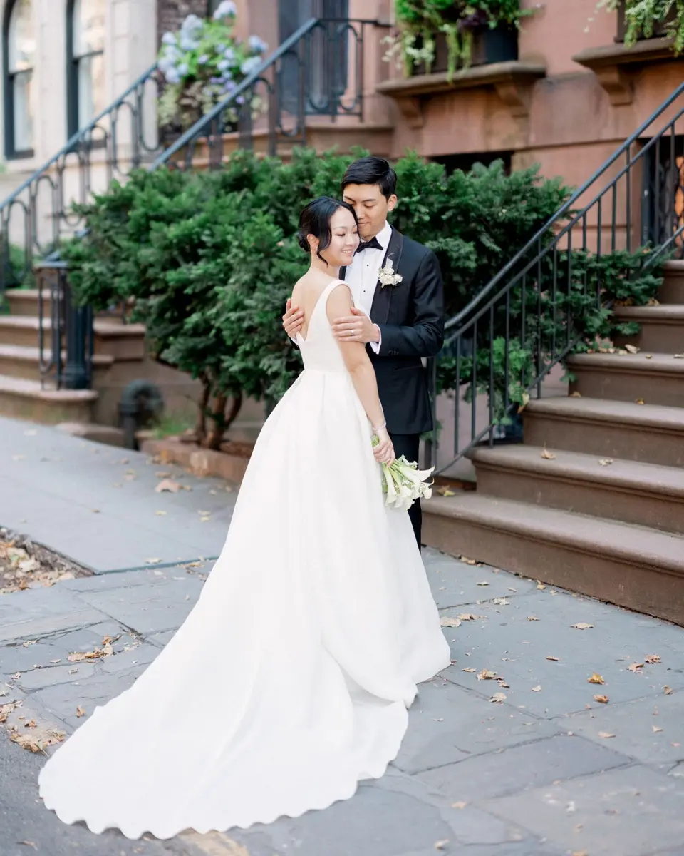 The groom rests his cheek against the bride's head as she looks down, smiling, her long train spread across the pavement in front of a Brooklyn brownstone with iron railings.