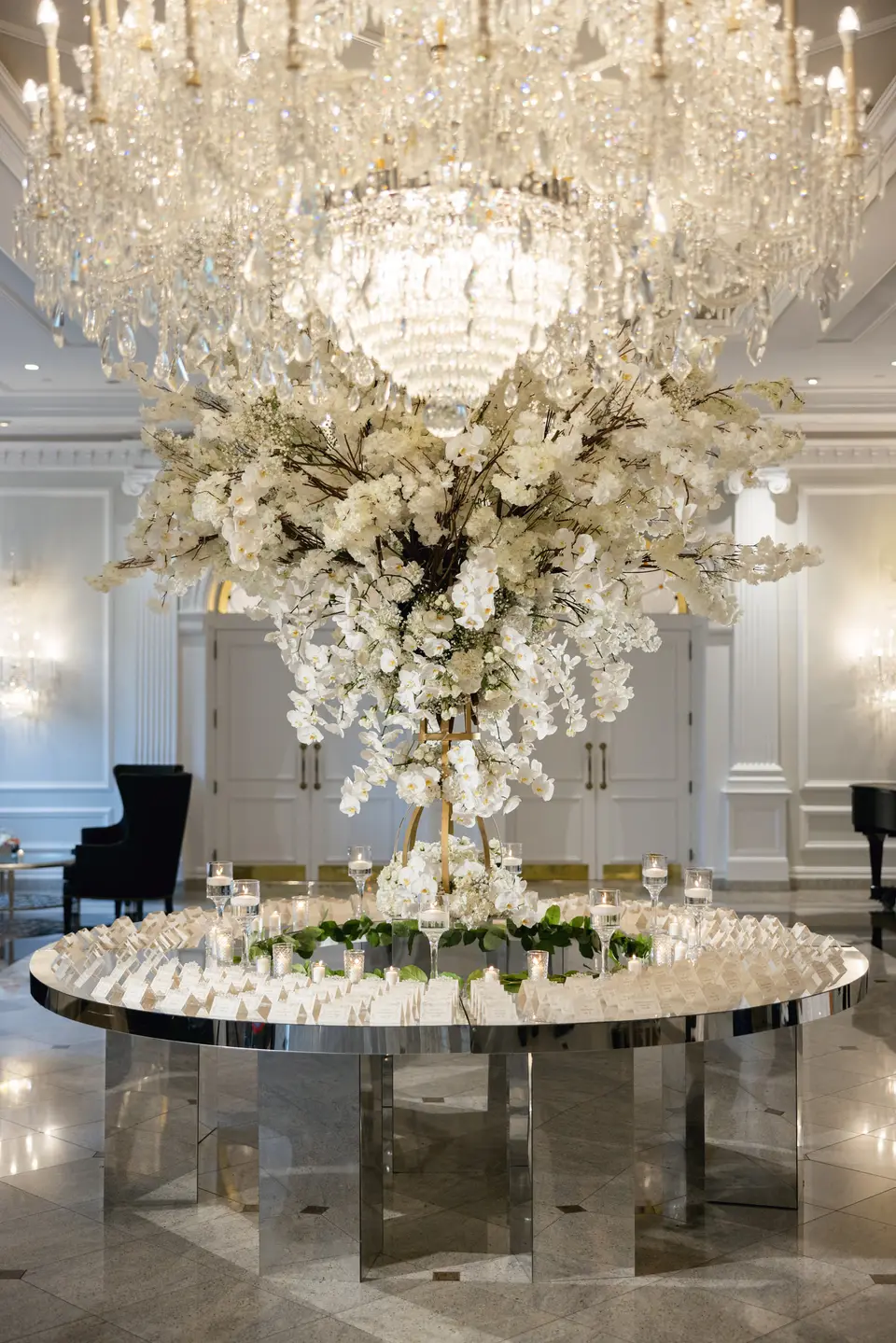 A round mirrored table of escort cards beneath a tall arrangement of white blossom and a vast crystal chandelier.