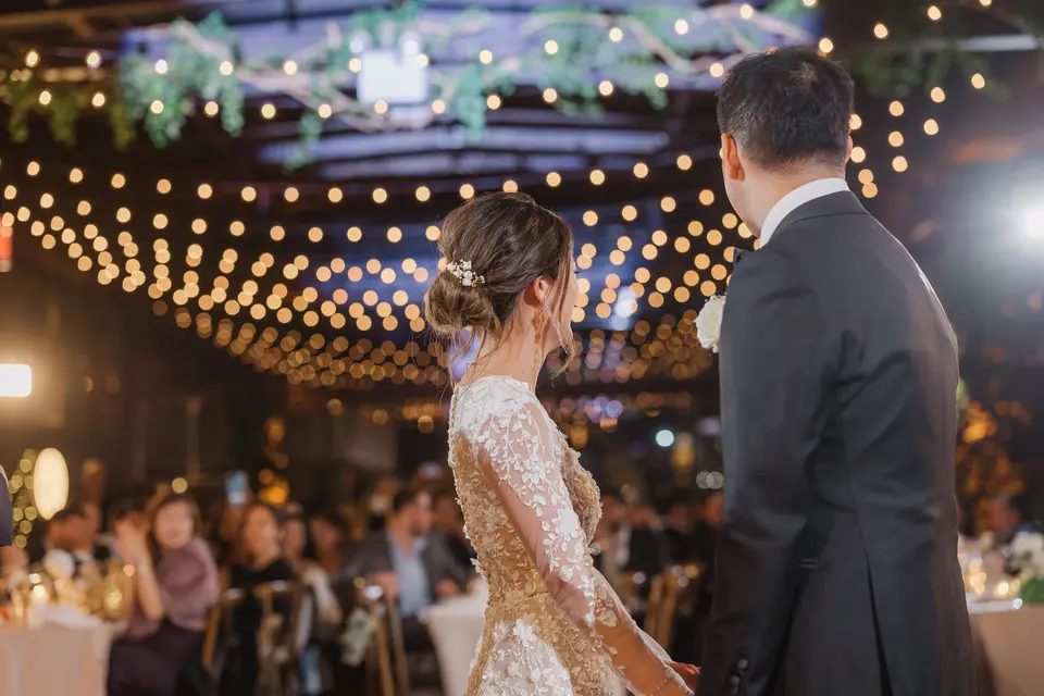 The couple face a room of guests beneath strings of warm lights and hanging greenery.