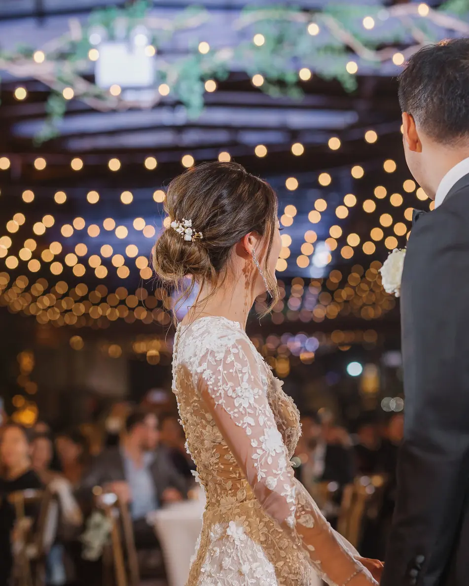 The couple face a room of guests beneath strings of warm lights and hanging greenery.