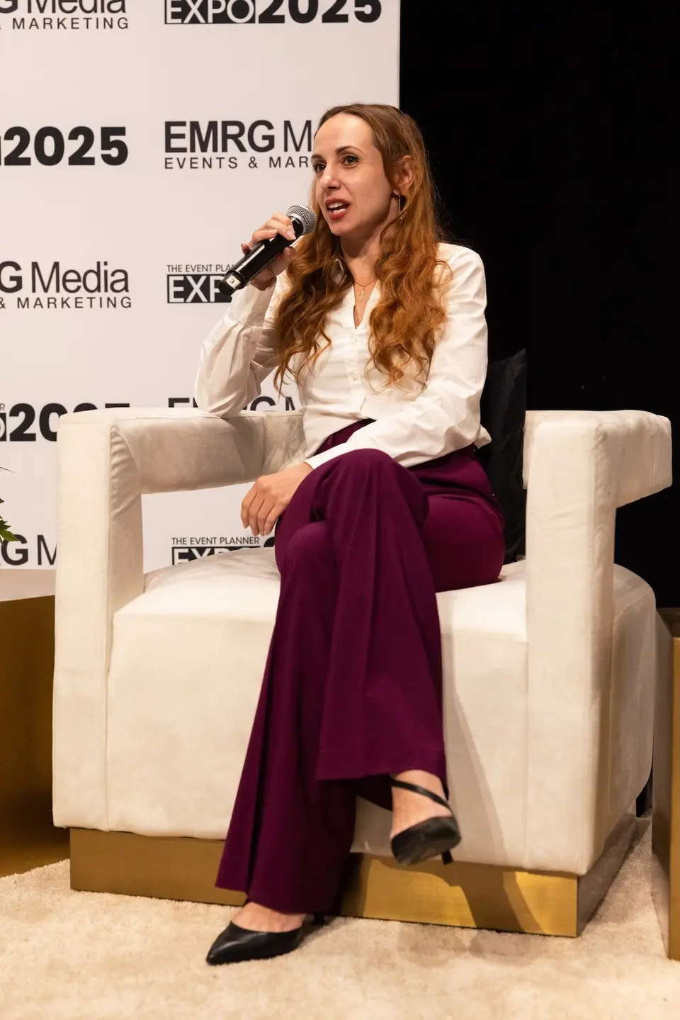 Olha Barabash speaks into a microphone from a white armchair on stage at The Event Planner Expo 2025, the Expo's logos behind her.