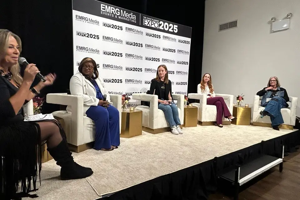A panel of five speakers in white armchairs on the Expo stage, a moderator speaking into a microphone.