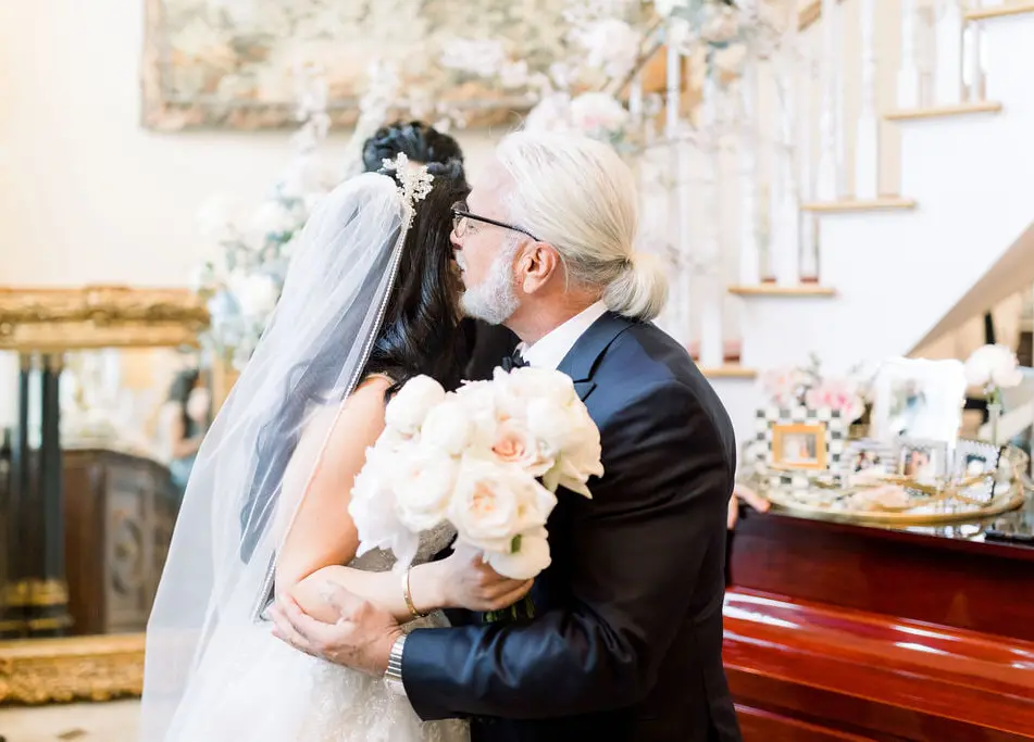 At the family home, a man with white hair kisses the bride on the cheek as she holds her bouquet of white roses.