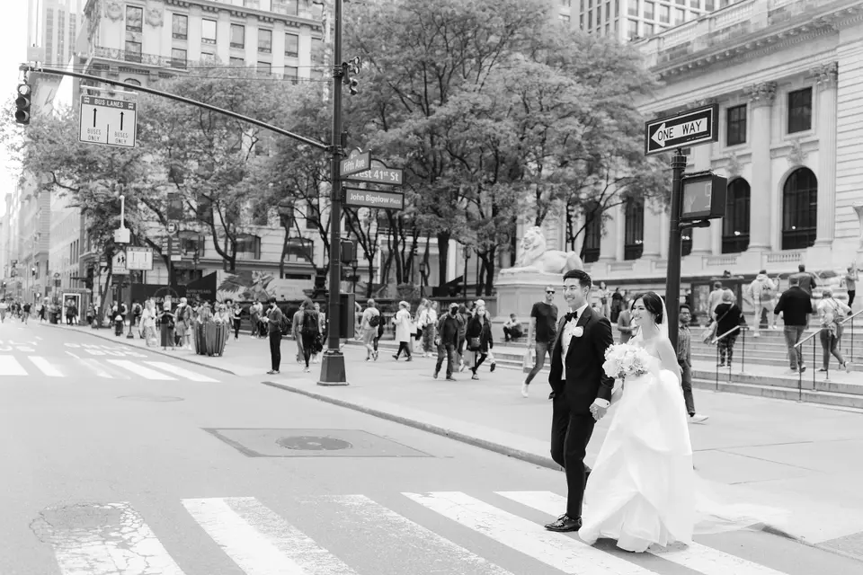 In black and white, the couple cross Fifth Avenue beside the New York Public Library, the city moving around them.