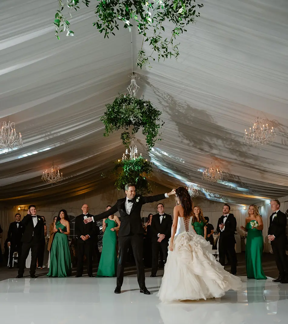 The couple's first dance on a white floor in the tent, guests watching beneath draped white fabric and hanging greenery.