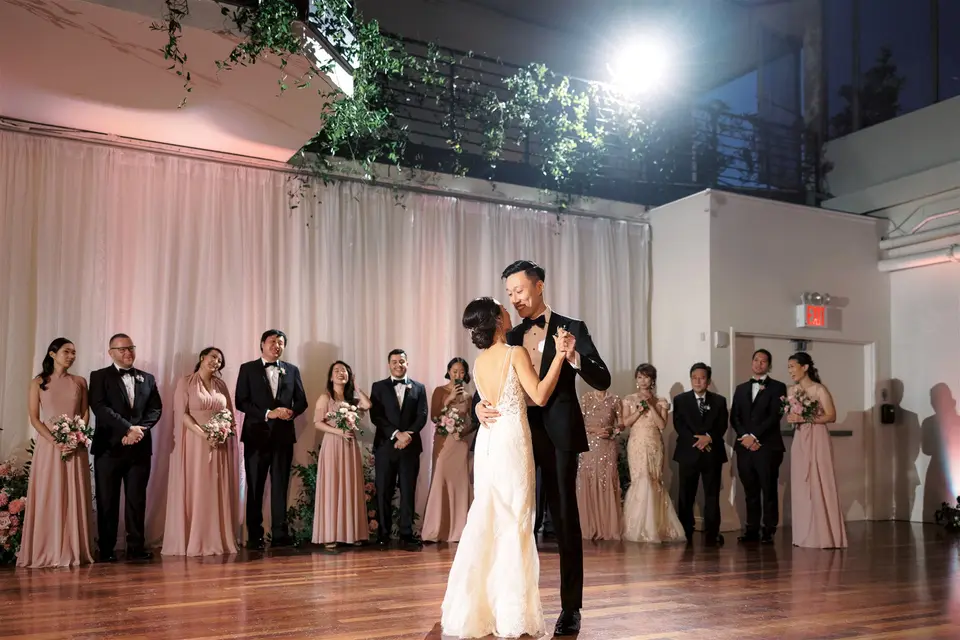 The couple dance on a wooden floor, the wedding party watching from the edge of the room in pink and black.