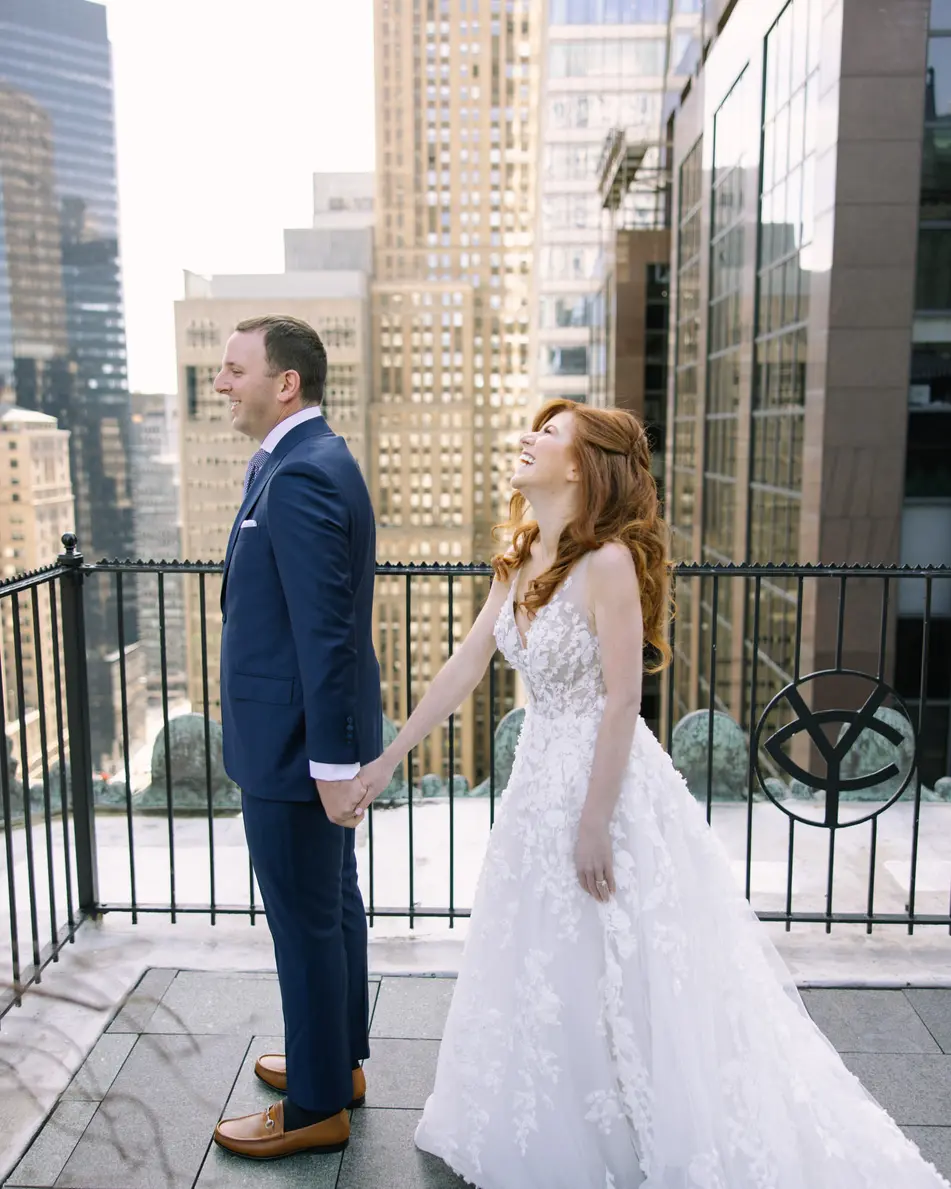 On the roof terrace high above Midtown, the bride laughs as she takes the groom's hand, the Yale emblem on the railing beside them.