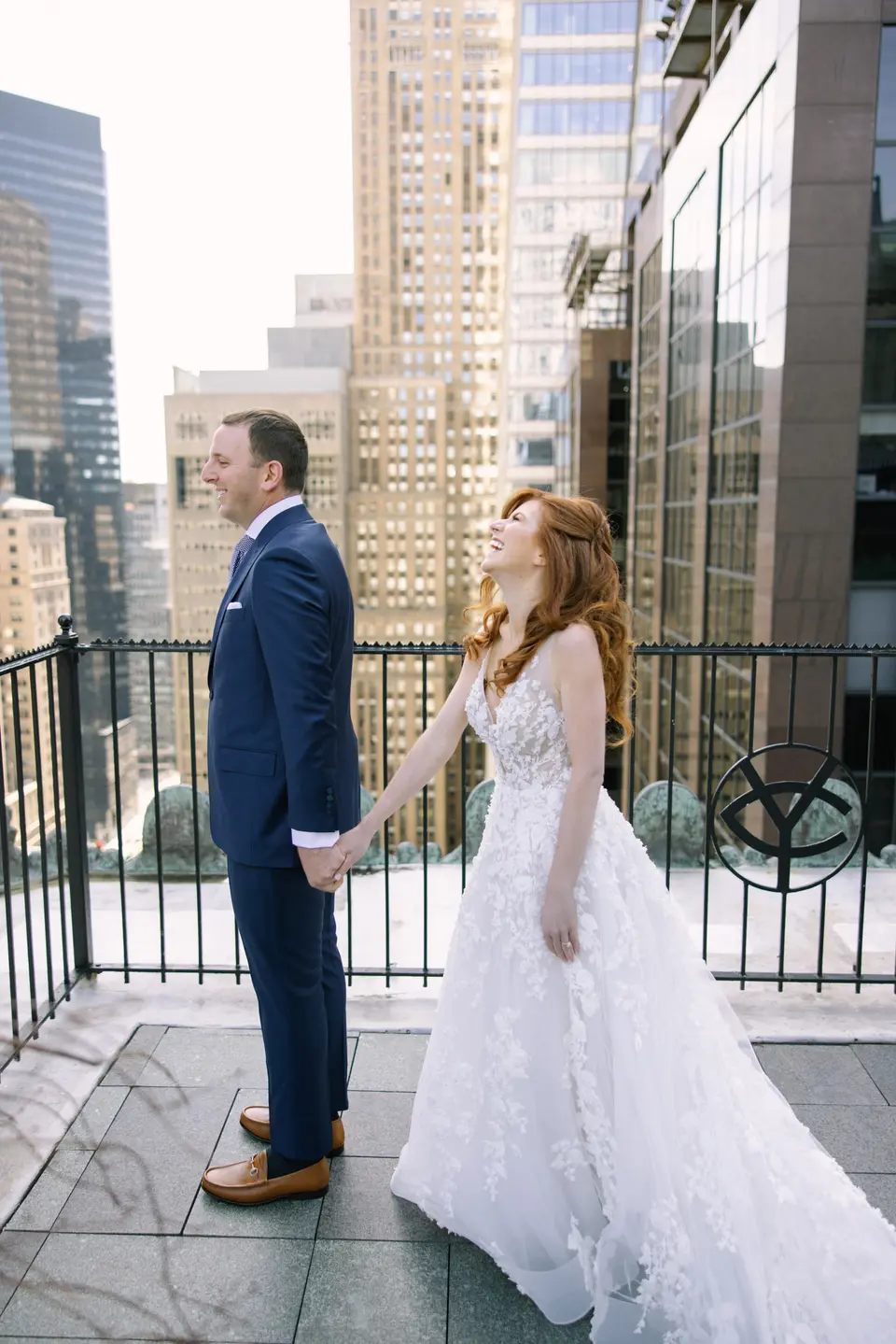On the roof terrace high above Midtown, the bride laughs as she takes the groom's hand, the Yale emblem on the railing beside them.