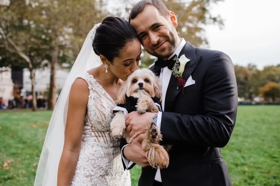 A bride and groom hold their small fluffy dog between them on a lawn, the bride kissing its head.