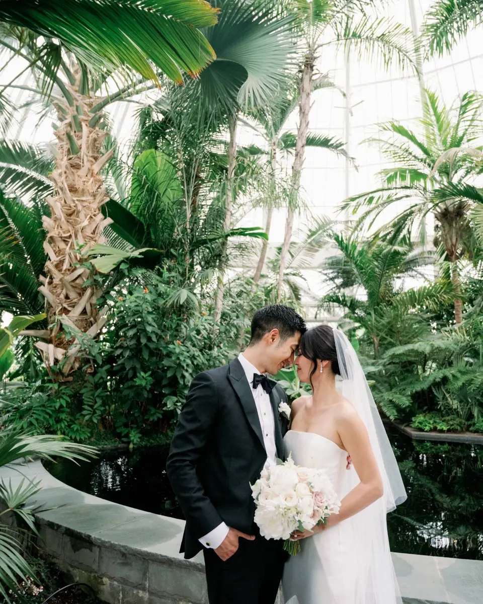 The bride and groom touch foreheads among tall palms and ferns inside the glasshouse at The New York Botanical Garden, her veil falling behind her.