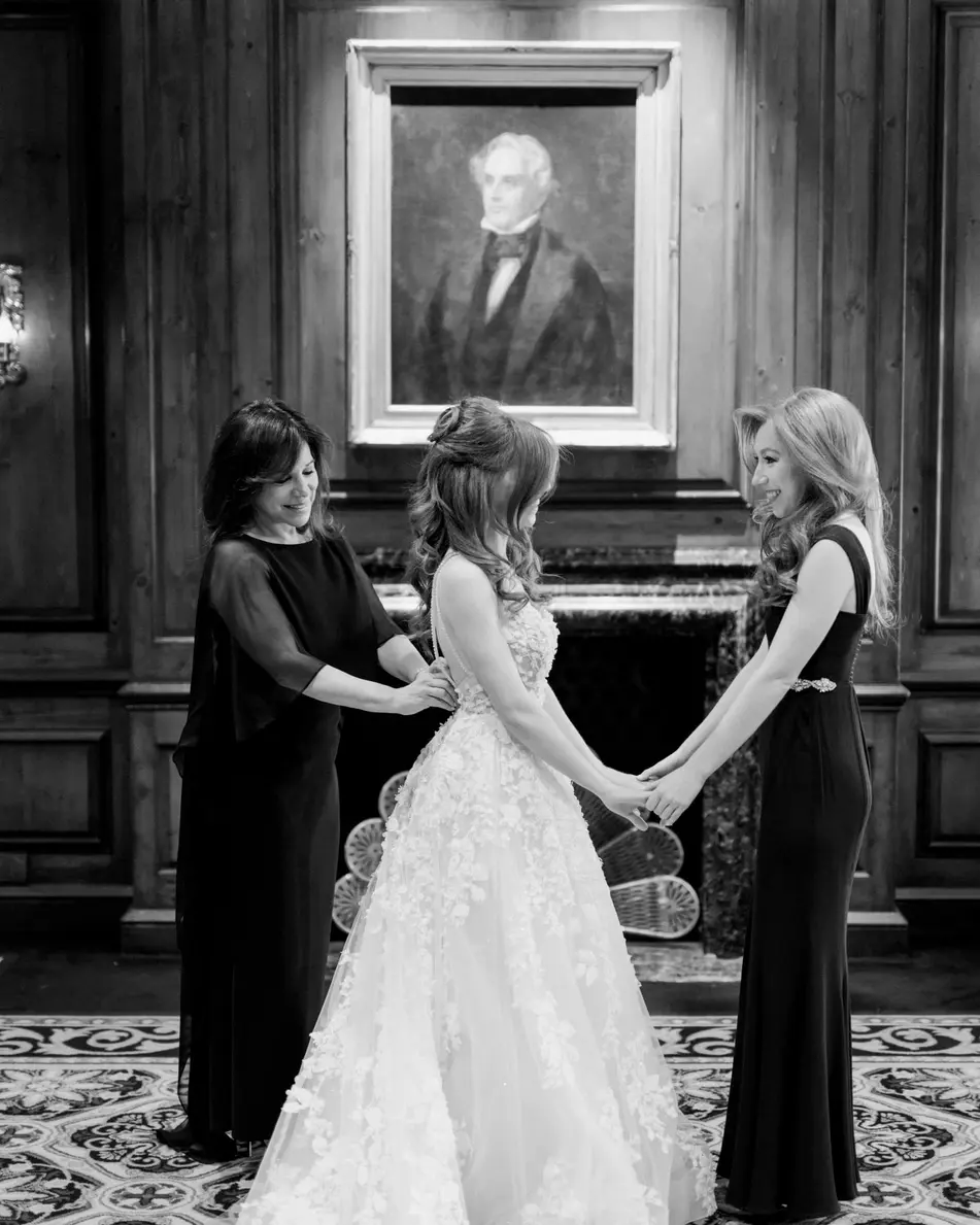 In a wood-panelled room beneath an old portrait, two women hold the bride's hands as she gets ready. Black and white.