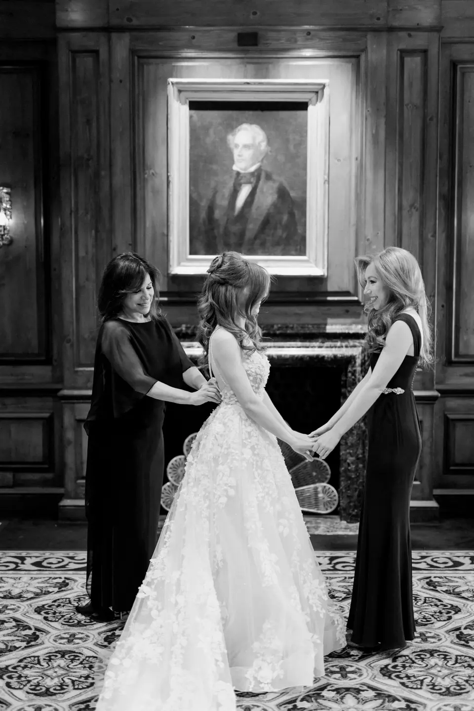 In a wood-panelled room beneath an old portrait, two women hold the bride's hands as she gets ready. Black and white.
