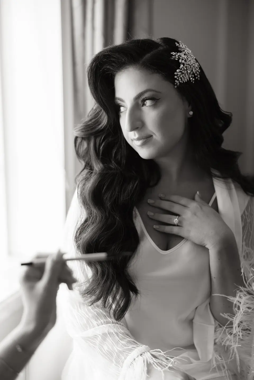 A makeup artist's brush at the bride's cheek as she looks toward the window, a hand at her collar. Black and white.
