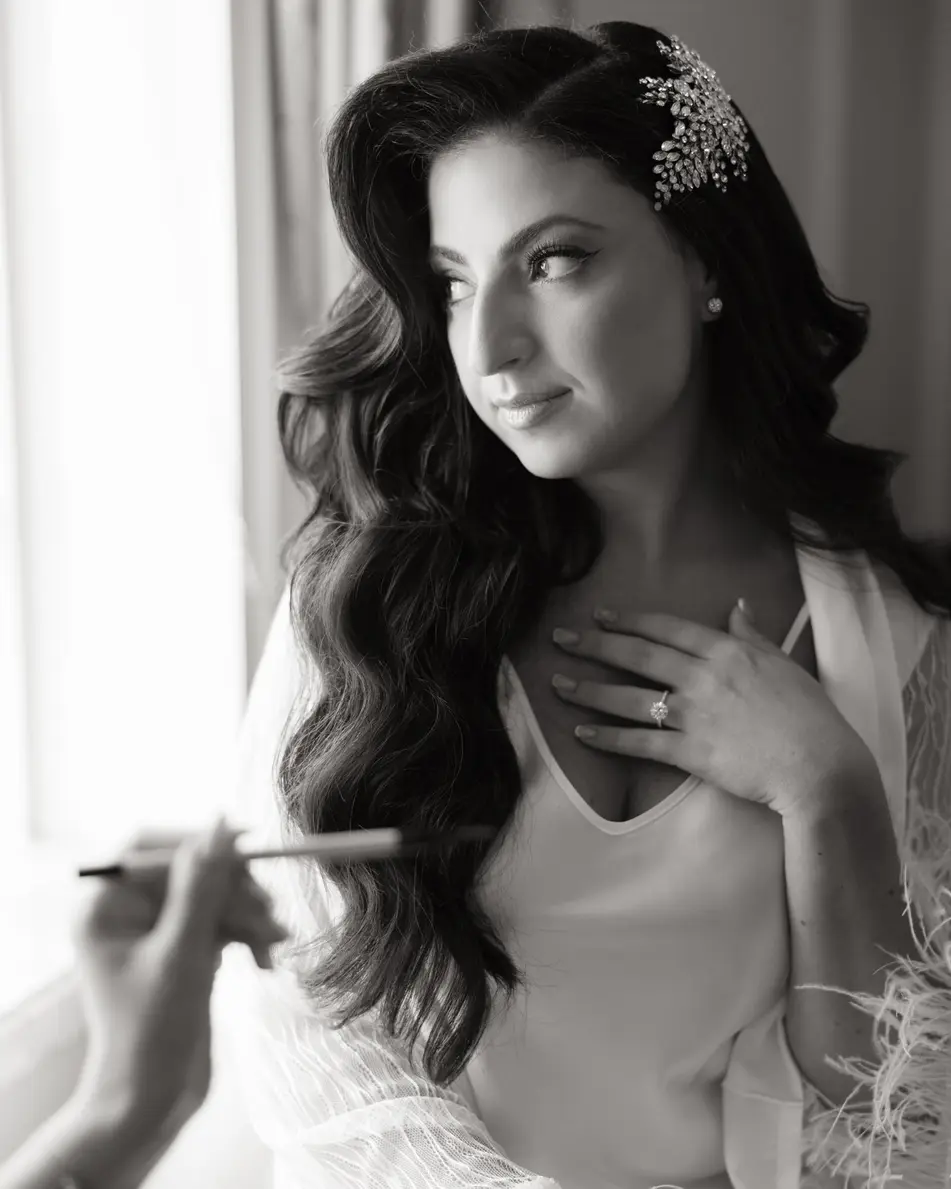 A makeup artist's brush at the bride's cheek as she looks toward the window, a hand at her collar. Black and white.