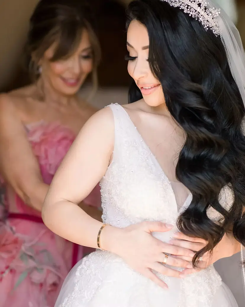 A woman in a pink dress helps the bride with her gown, both of them smiling, the bride’s dark hair over one shoulder.