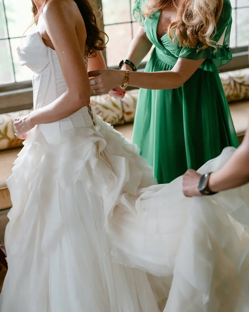 A bridesmaid in an emerald dress fastens the back of the bride's gown by the window.