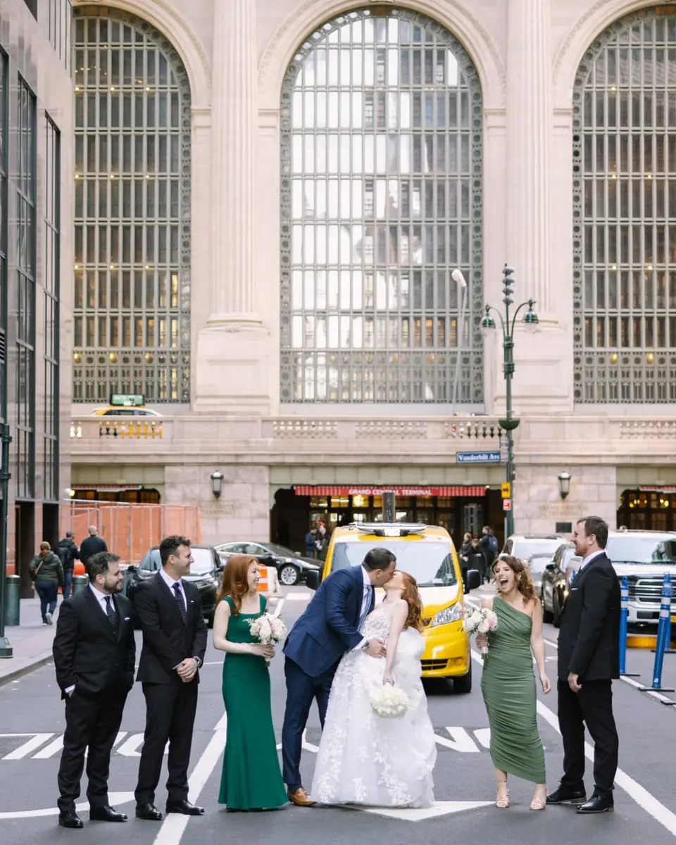 The couple kiss in the middle of Vanderbilt Avenue, their wedding party lined up beside them in black suits and green dresses, the tall arched windows of Grand Central behind.
