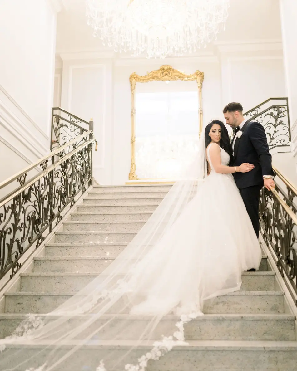 The bride and groom stand on the white marble staircase at The Rockleigh, her cathedral veil cascading down the steps.