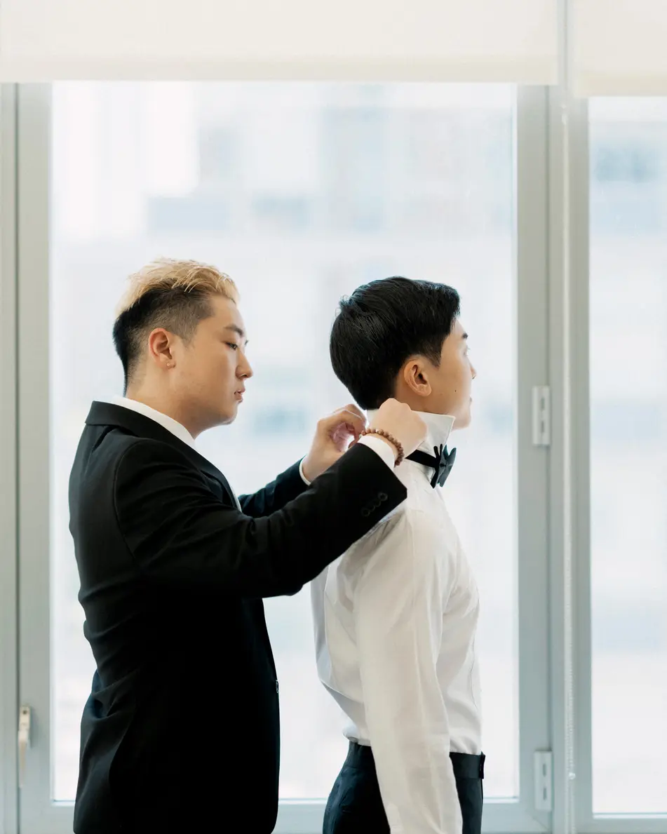 By a bright window, a friend adjusts the groom's bow tie.