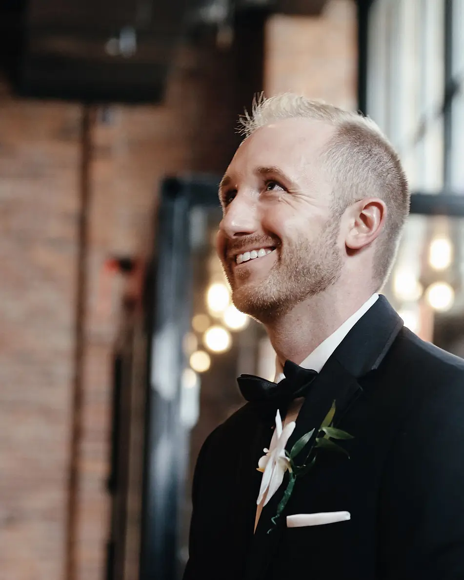 The groom smiles as he waits in a room of brick and tall windows.
