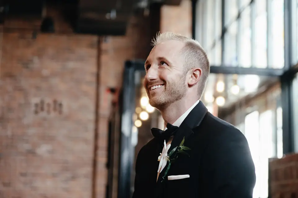 The groom smiles as he waits in a room of brick and tall windows.
