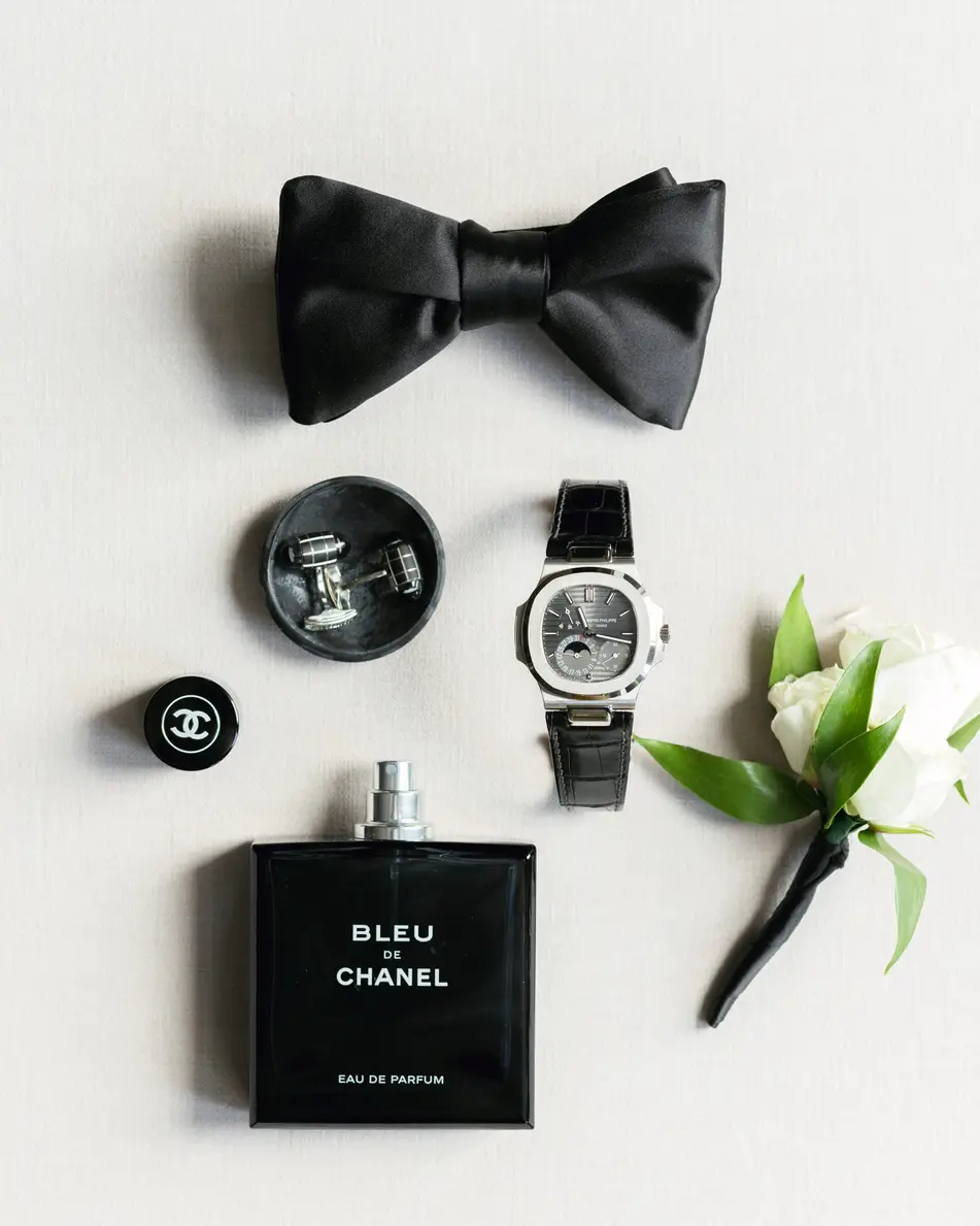 A black bow tie, cufflinks, a watch, cologne and a white boutonniere laid out on white.