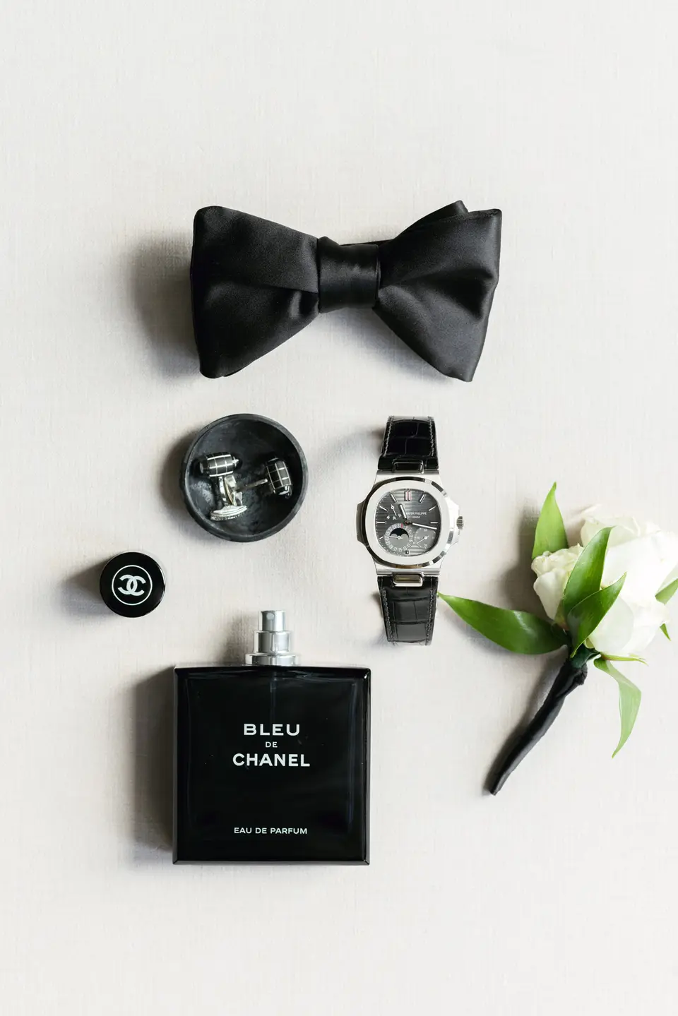 A black bow tie, cufflinks, a watch, cologne and a white boutonniere laid out on white.