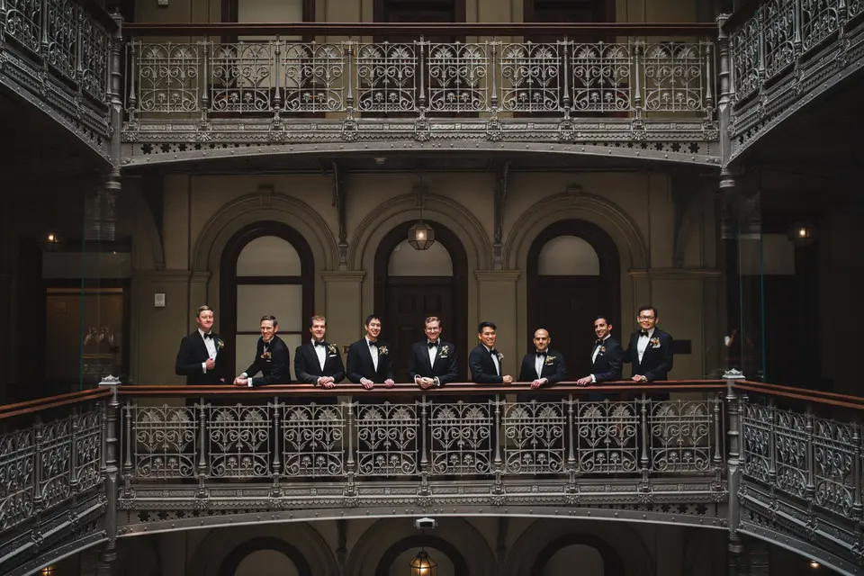 The groom and his groomsmen line an ornate iron balcony in a tall Victorian atrium.