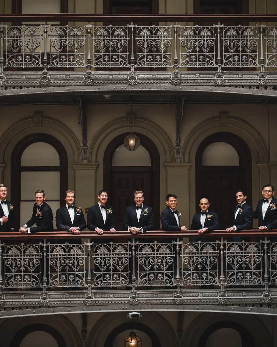 The groom and his groomsmen line an ornate iron balcony in a tall Victorian atrium.